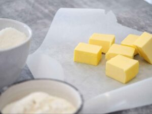 Cubes of yellow butter on parchment paper next to bowls of flour or sugar.