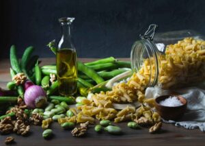 A variety of food ingredients including pasta, olive oil, green beans, walnuts, and an onion on a wooden surface.